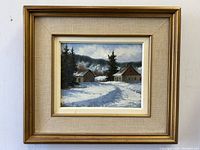 Frontal view of the framed oil painting showing a snowy countryside scene with old houses and pine trees.