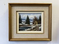 Frontal view of the framed winterscape oil painting showing the cabin, trees, snow, and water.
