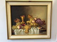 Frontal image of the large still-life oil painting depicting grapes, peaches, a brass vase, and a glass of wine on a draped tablecloth in a gold frame with cream mat.