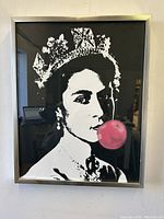 Framed black and white print of Queen Elizabeth II blowing pink bubble gum against black background, hung on wall.