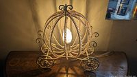 Lamp designed as Cinderella style carriage with pink beads around circular metal frame, lit bulb visible inside, placed on wooden surface.