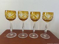 Four vintage amber-colored Bohemian etched glass wine goblets with clear faceted stems and ribbed bases, arranged side by side on a wooden surface against a white background.