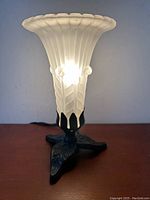 Front view of the lamp illuminated on a wooden surface. Shows trumpet shaped frosted glass shade and black metal leaf base.