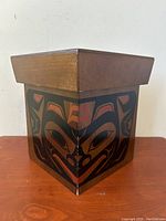Side angle view of wooden rectangular storage box with detailed Haida style painting in black and red and natural wood top edge.