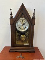 Front view of vintage Jupiter steeple mantle clock, showing white dial with Roman numerals, wooden pointed case, glass front with gold designs, brass pendulum, and winding key on table.