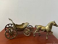 Side view of the solid brass horse attached to the wheeled carriage on a wooden surface showing the carriage's scrollwork detail and the horse's prancing pose.