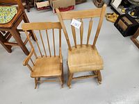 Photo of two children's wooden rocking chairs, one with armrests and one without, placed side by side showing worn natural wood finish.