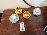 Overview of 4 cups with their matching saucers, arranged on a wooden surface showing variety of ceramic and metallic styles.