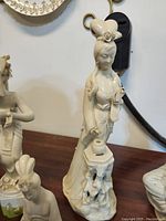 Close up of a ceramic figurine of a Chinese woman holding a flower near a pedestal, showing delicate details in the sculpture.