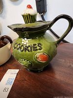 Green ceramic cookie jar with colorful decorative accents and cupcake-shaped lid, set on a wooden surface.