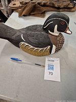 Wooden duck decoy painted with naturalistic colors and fine feather detail, placed on a table with a pen and auction lot card.