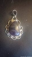 Close-up photo of a teardrop-shaped amethyst pendant in a silver filigree setting on dark textured surface.