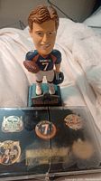 Front angle showing John Elway bobblehead in Broncos uniform on Class of 2004 base with 5-pin set in black case below it.