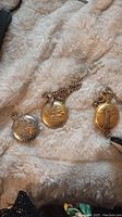 Three pocket watches, silver-tone with bird design, gold-tone with ship design and chain, gold-tone with figure design and chain laid out on a beige plush fabric.