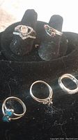 Five silver-tone rings displayed on black velvet stand, showing variety of styles including heart, wedding set, mothers ring.
