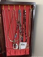 Nine necklaces hanging on hooks inside a case with maroon backing. Varied styles including beaded, metallic, and chain. Pendants included on some.