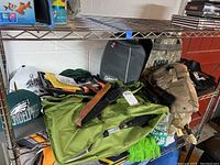 Metal shelf holding multiple camping items including green Camelbak pack, Coleman carrying case, and beige backpack.