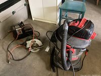 Photo showing Black & Decker circular saw with orange cord and black and red wet/dry shop vacuum with hose attached, both placed on the floor.