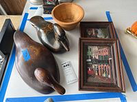 Two carved wooden duck decoys, two framed relief artwork pieces, and a wooden bowl arranged on a table with blue tape border. Shows size and condition.