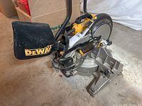 Side view of DeWalt radial arm saw on floor showing blade, dust bag, handle, and part of the saw base.