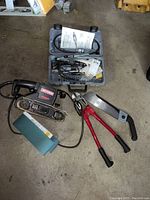 Craftsman electric belt sander with sanding belt attached lying on concrete floor, alongside bolt cutters and hand saw.