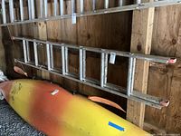 Photo showing entire aluminum extension ladder leaning against wooden wall above a kayak inside a garage.