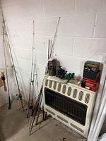 Eleven rods leaning against wall next to heater with loose reels and game camera visible