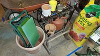 Overview photo showing several plant pots, a green folding garden stool inside a large pot, a black decorative metal plant stand, and a wooden frame basket/tray on the floor.