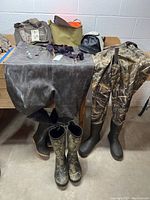 Photo showing two pairs of hunting waders laid out on a table with attached boots and additional accessories including bags, suspenders, and duck call arranged around them.