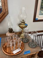 Full lot showing two oil lamps, two pewter hurricane candle holders, six vintage drinking glasses with gold leaf designs, and copper tray on table.