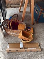 View of four Longaberger baskets of different sizes and two other smaller woven baskets, with IKEA coat hooks in original packaging in front of them on the floor in a garage.