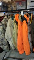 Photo of multiple men's hunting coats and jackets hanging on rack including camouflage Cabela's coat and blaze orange jackets.