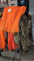 Multiple men's hunting vests and pants hanging on a rack including orange high-visibility and camouflage patterns