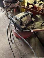 Full image showing red cylindrical air compressor with black top and handle, connected hose and power cord on concrete garage floor.