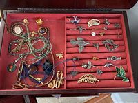 Top-down view of a jewelry box with various pins, brooches, necklaces, and rings on red felt