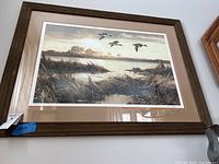 Framed print showing a marsh scene with ducks flying, wooden frame and glass covering. Photo taken to capture overall appearance and condition.