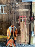 Three sleds displayed against a wooden wall: a tall black metal framed sled, a colorful plastic sled with orange and black flames, and a vintage Flexible Flyer wooden sled with red metal runners and rope.