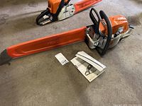 Chainsaw laid flat with orange bar scabbard and owner’s manual