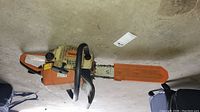 Side and top view of the Stihl 025 chainsaw lying on a concrete floor showing wear, dirt, and intact bar with chain and orange cover