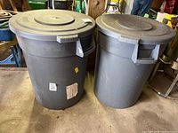 Photo showing two large gray plastic Carlisle trash cans side by side with matching lids, dirt and wear visible on surfaces.