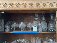 Shelf displaying multiple pieces of Waterford and cut glass including decanters, brandy glasses, a divided dish and a vase, arranged for photo.