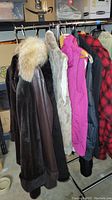 Dark fur coat with light fur collar and partial view of additional coats and vests on rack