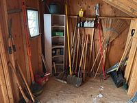 View of a corner with multiple yard tools including axes, shovels, a rake, pole clippers, fuel cans and a metal tool shelf.