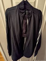 Front view of black Adidas women's jacket hanging, showing full zipper and white stripes on sleeves.