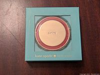 Front view of compact mirror in blue Kate Spade branded box, gold tone with red trim visible through window.