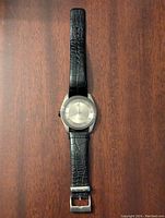 Front view of the Kenneth Cole watch showing the silver round face and black leather strap lying flat on a wooden surface.