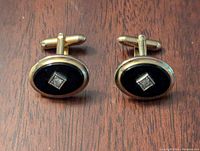 Pair of men's oval-shaped cufflinks with gold-tone surrounds and black enamel centers each featuring a diamond-shaped silver-tone accent, lying on wooden surface.