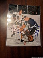 Full signed hockey photo showing Bobby Hull and Johnny Bower in action on ice, protected in plastic sleeve.