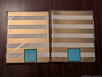 Two new Kate Spade notebooks with gold and white horizontal stripes, wrapped in plastic, placed side by side on a wooden surface.