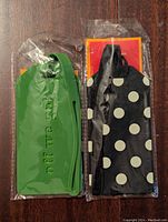 Front view of two luggage tags sealed in original plastic packaging; green one embossed with 'Off We Go' text and black one with white polka dots.